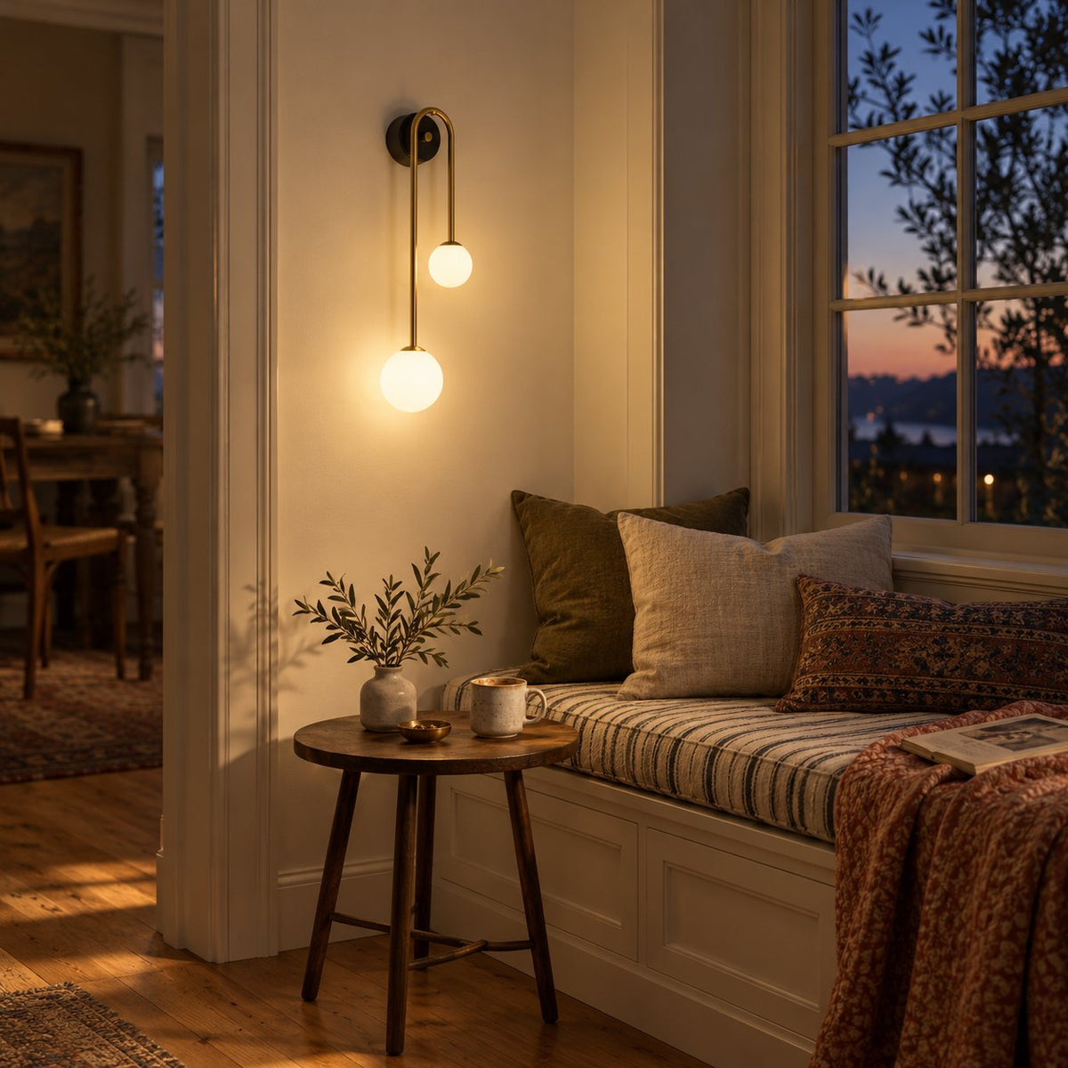 Alcoutim arc wall sconce with two lit opal glass globes beside a cushioned window seat at dusk.