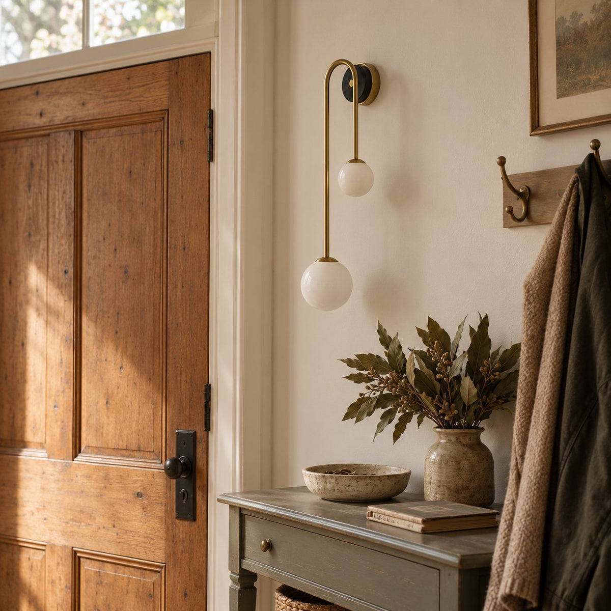 Alcoutim arc wall sconce with two lit opal glass globes beside a wood entry door and coat hooks.