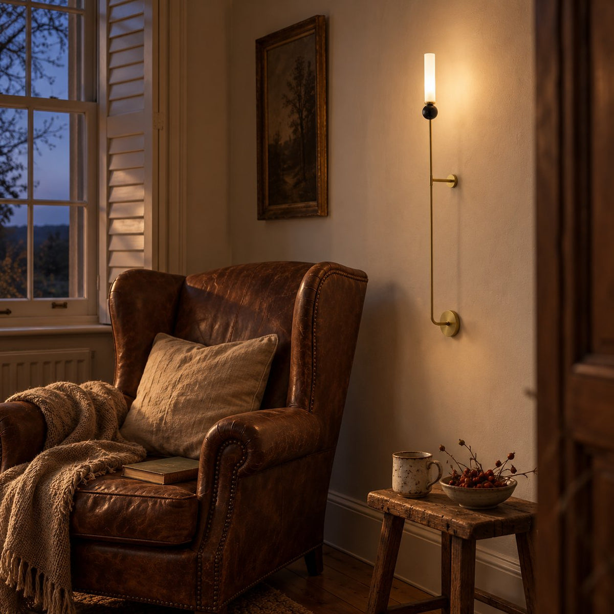 Aljezur vertical marble wall sconce lit on brass beside a leather wingback chair at dusk.