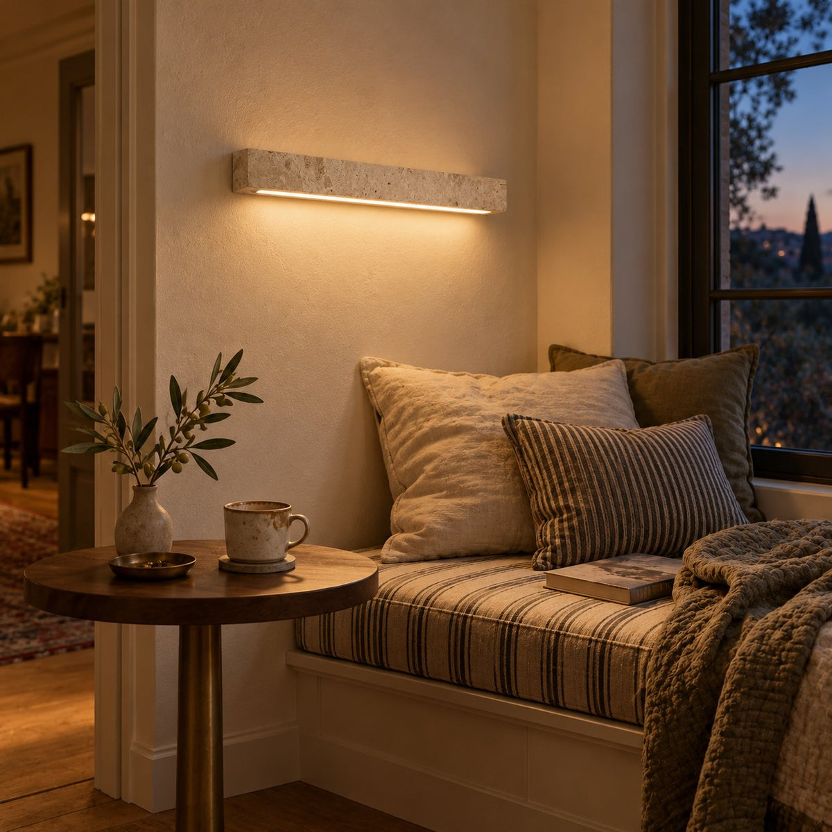 Barrancos travertine linear sconce glowing warmly above a cushioned window reading nook.