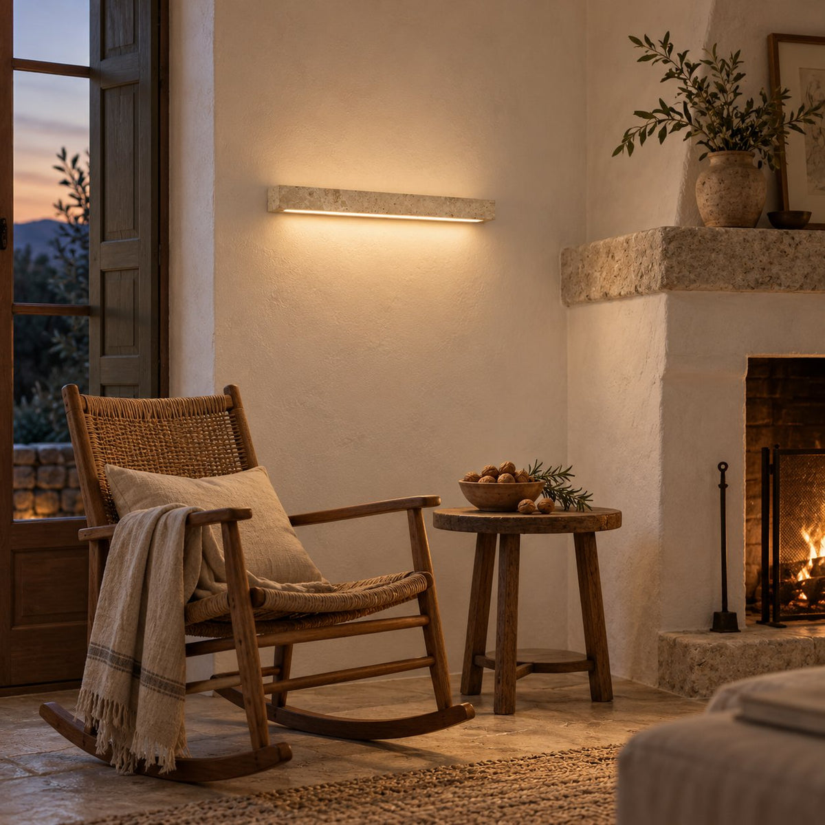 Barrancos linear wall sconce in cream travertine, lit beside a rocking chair and stone fireplace.