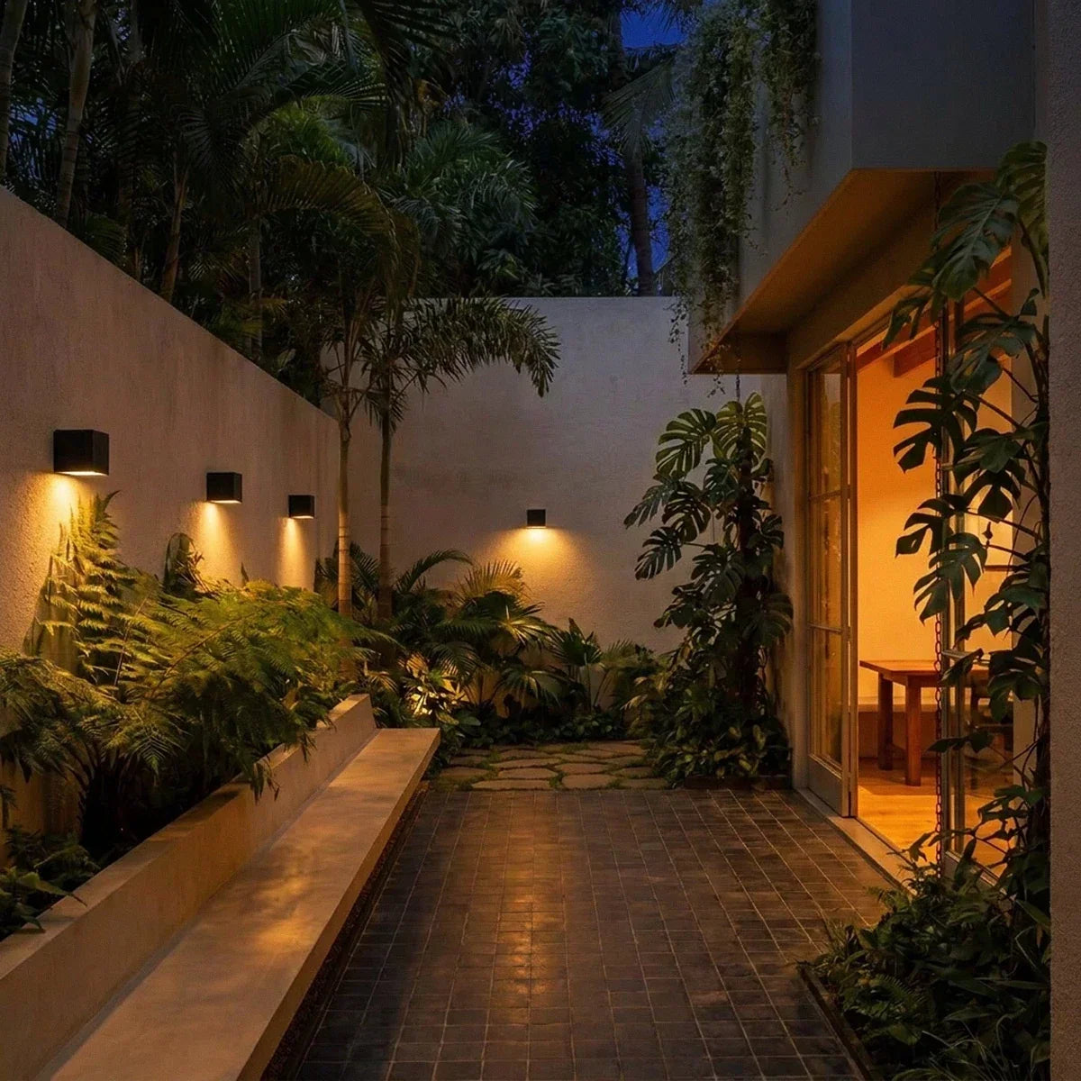 Same garden courtyard at dusk with the Clérachic black cube wall lights glowing warm down the wall beside a lit interior.
