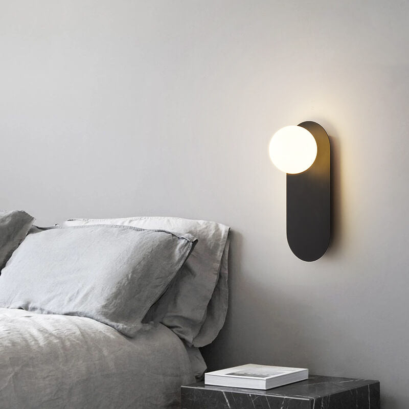 Valença black wall sconce lit on a gray bedroom wall above a black marble nightstand beside a bed