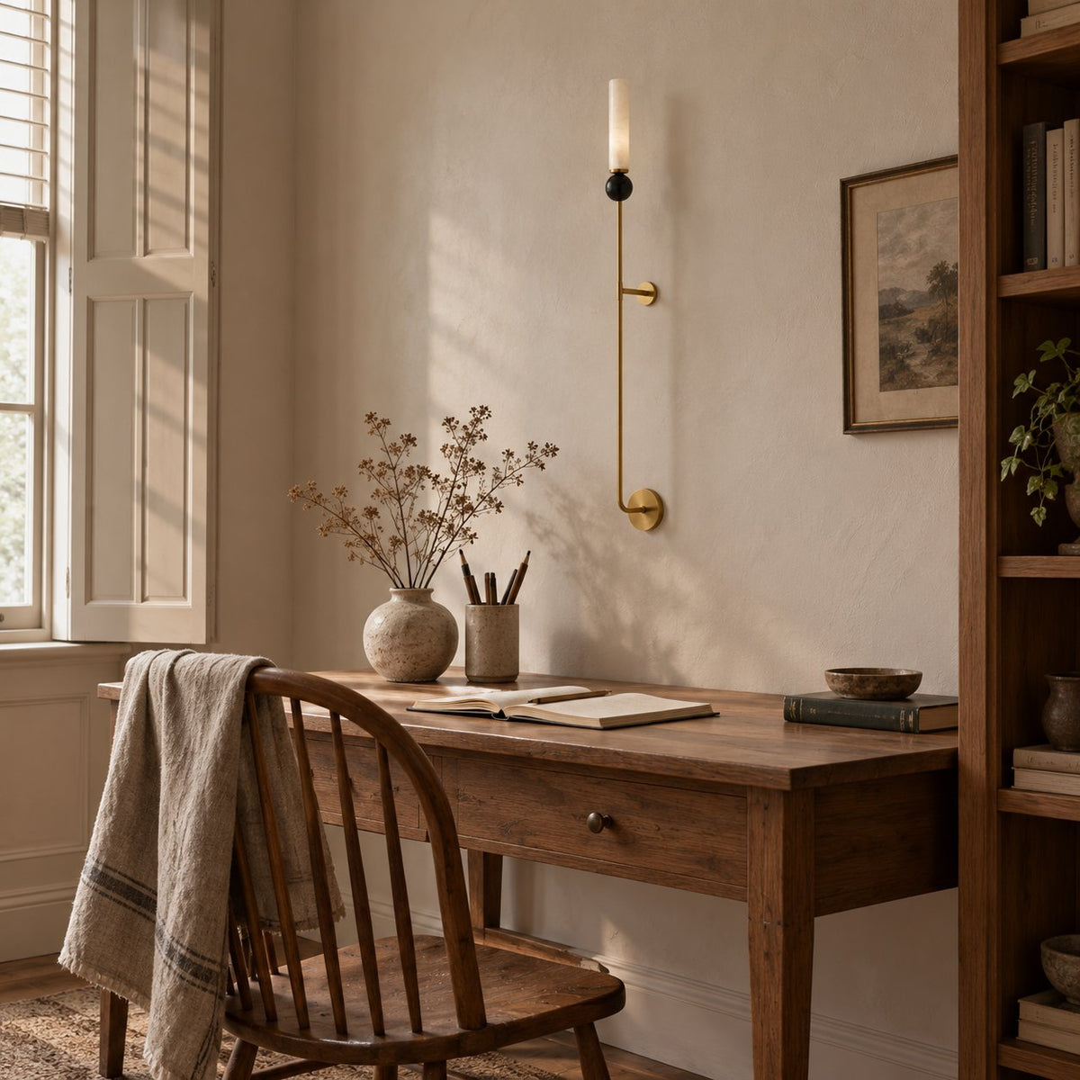 Aljezur vertical marble wall sconce on brass above a wood writing desk by a sunlit window.