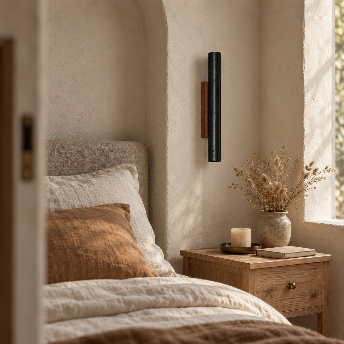 Arraiolos stone column sconce, black with a walnut mount, lit in an arched bedroom alcove.