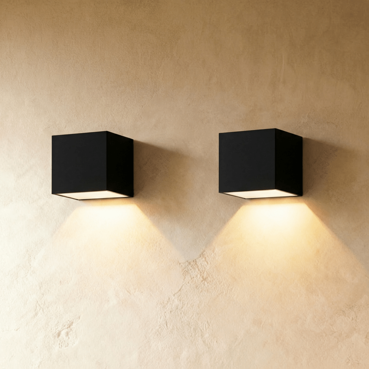 Pair of Clérachic black cube wall lights side by side on limewash plaster, each throwing a warm cone of light downward.