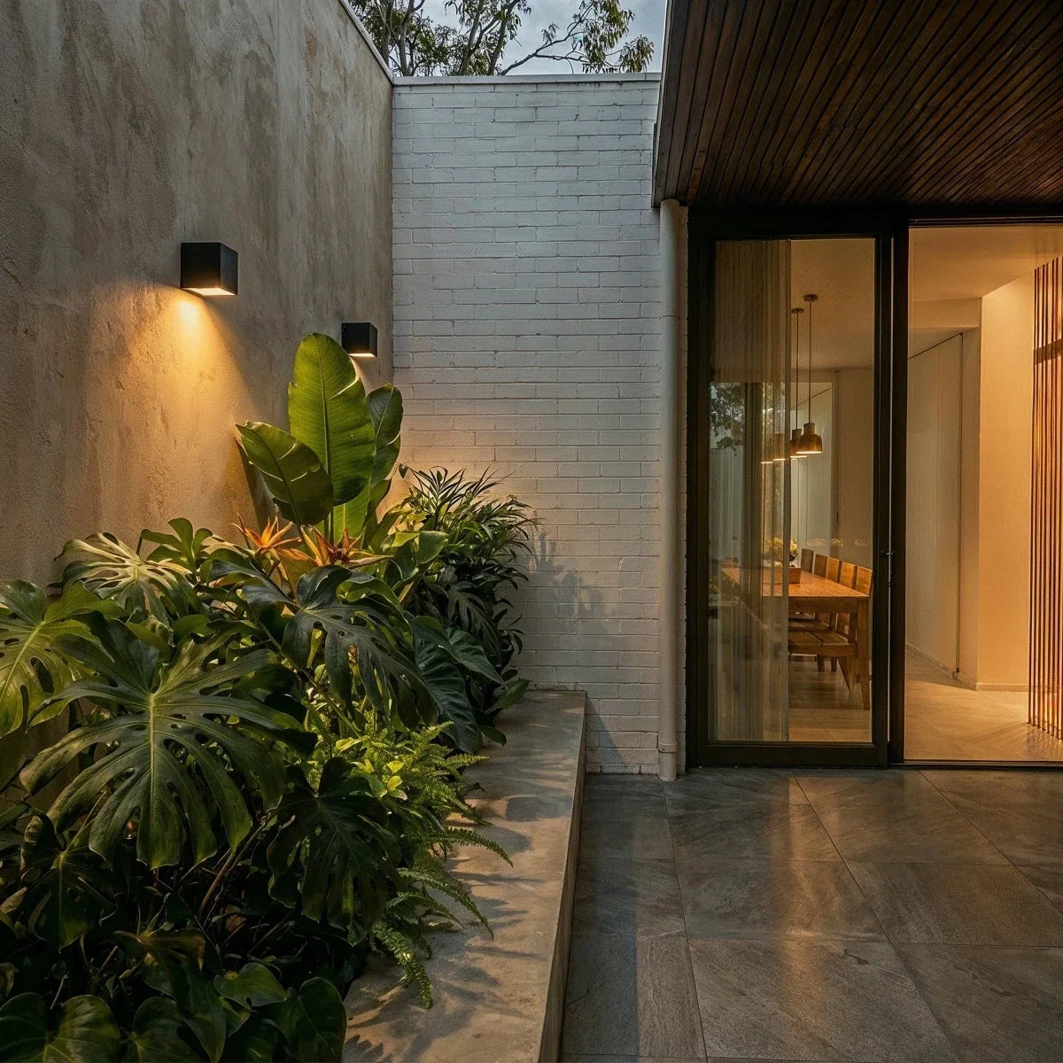Clérachic black cube wall lights on a courtyard wall at dusk beside a glazed door, with the room inside glowing warm.