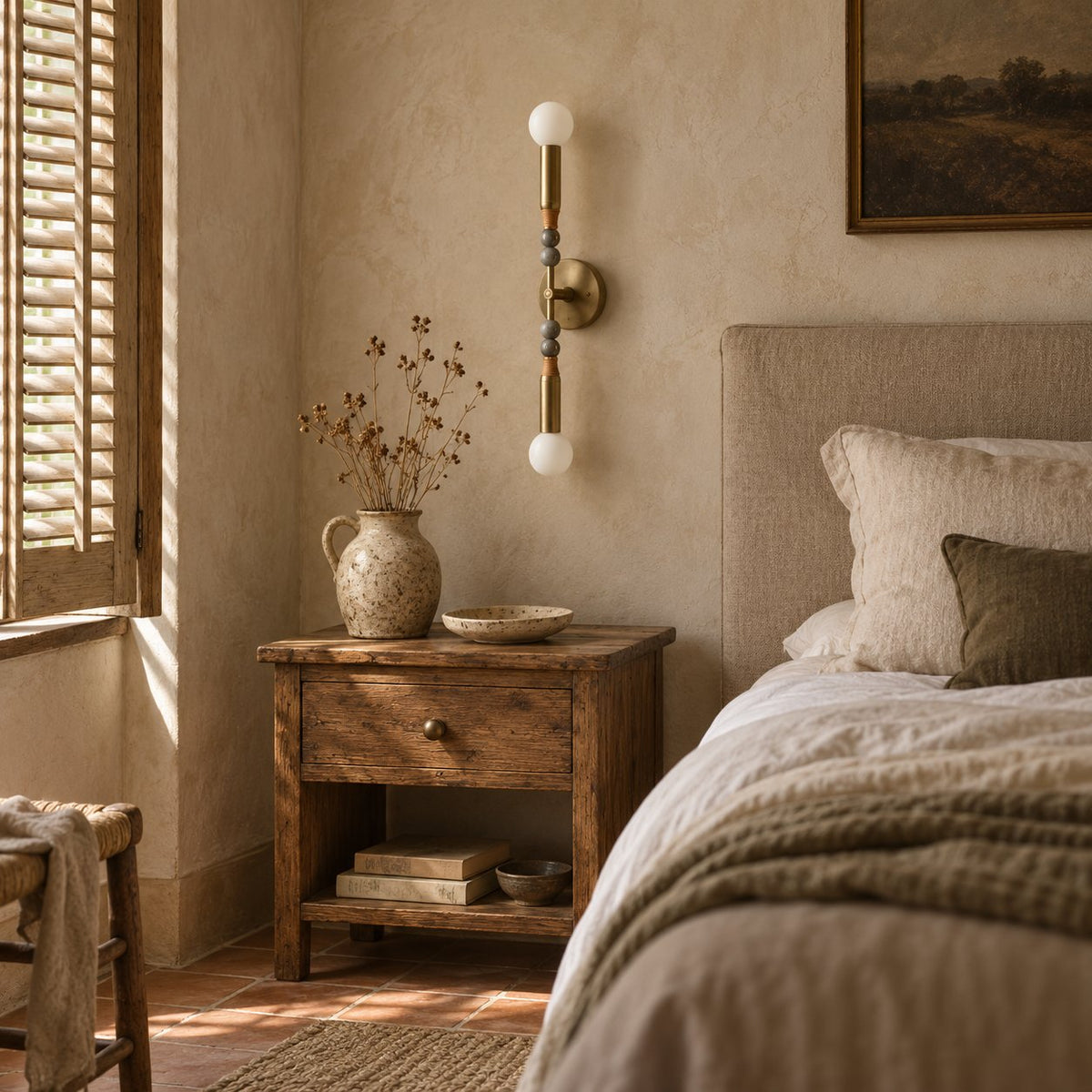 Torto brass double-globe wall sconce above a rustic nightstand with a speckled stoneware jug.